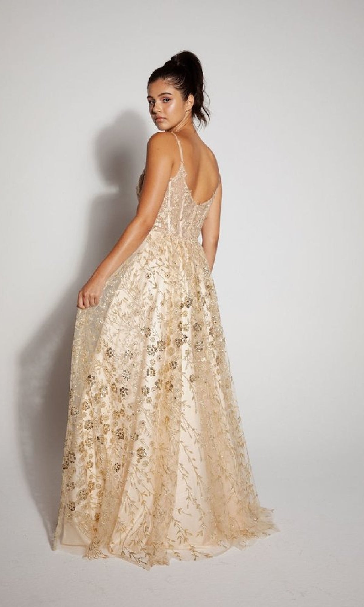 A woman wears the Long Glitter A-Line Prom Dress: Eureka 9908, featuring gold embroidery and a sleeveless, floor-length design, as she poses against a plain white background looking over her shoulder.